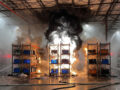 Warehouse storage racks engulfed in flames with thick black smoke rising between shelves and blue/orange containers on fire; hoses lie on the wet floor.