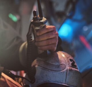 Emergency responder grips a radio microphone attached to a helmet in a dim, smoky environment.