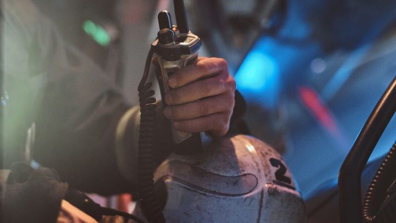 Emergency responder grips a radio microphone attached to a helmet in a dim, smoky environment.