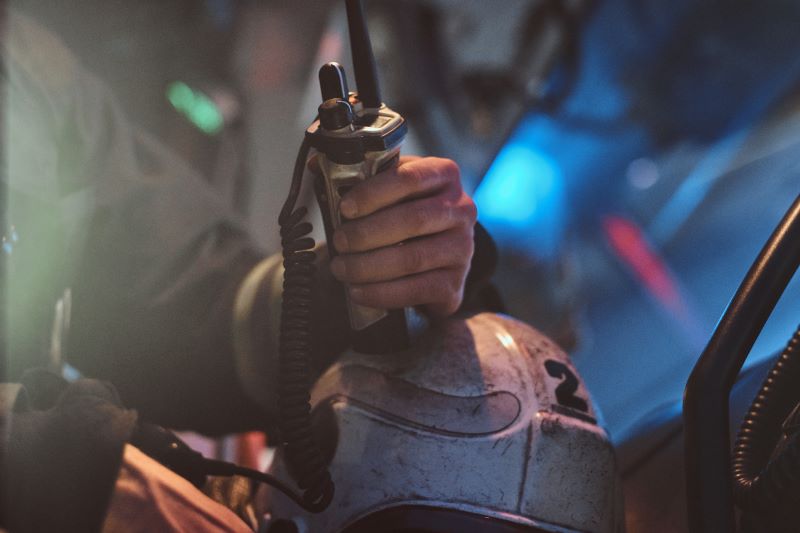 Emergency responder grips a radio microphone attached to a helmet in a dim, smoky environment.